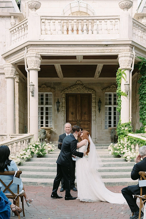 Wedding kiss as bride in veil and strapless lace dress embraces groom in black tuxedo before stone staircase mansion ceremony backdrop