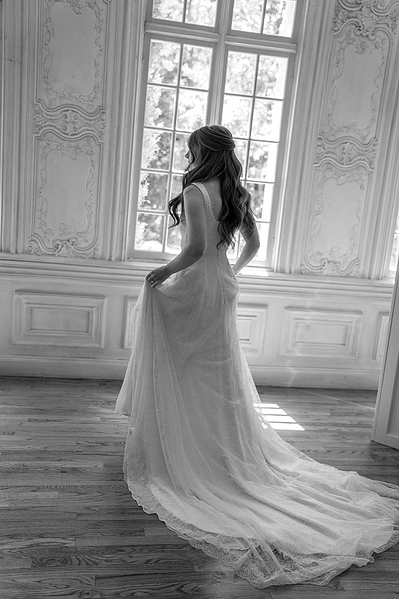 Bridal portrait in black and white of a bride by window, showing a lace train on her wedding dress in an ornate, sunlit room