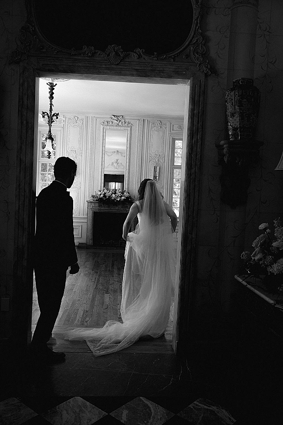 Wedding first look as the bride enters the room, long veil trailing behind, while the groom in a black suit watches near an ornate mirror