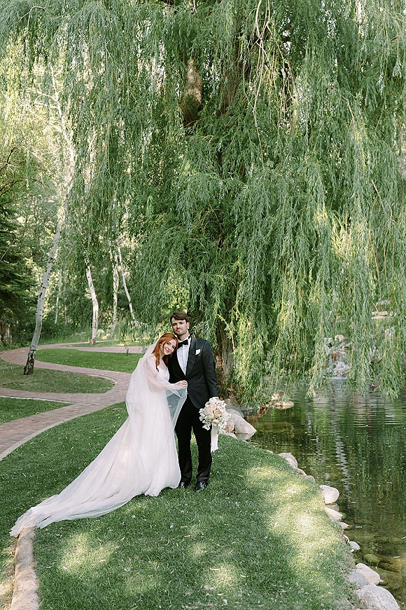 Couple portrait of bride leaning on groom, holding a blush rose bouquet with a long veil by a willow tree near a pond