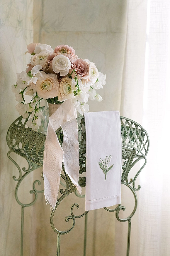 Wedding bouquet of blush and white blooms in a glass vase with ribbon, styled on a wrought iron chair by sheer curtains in window light