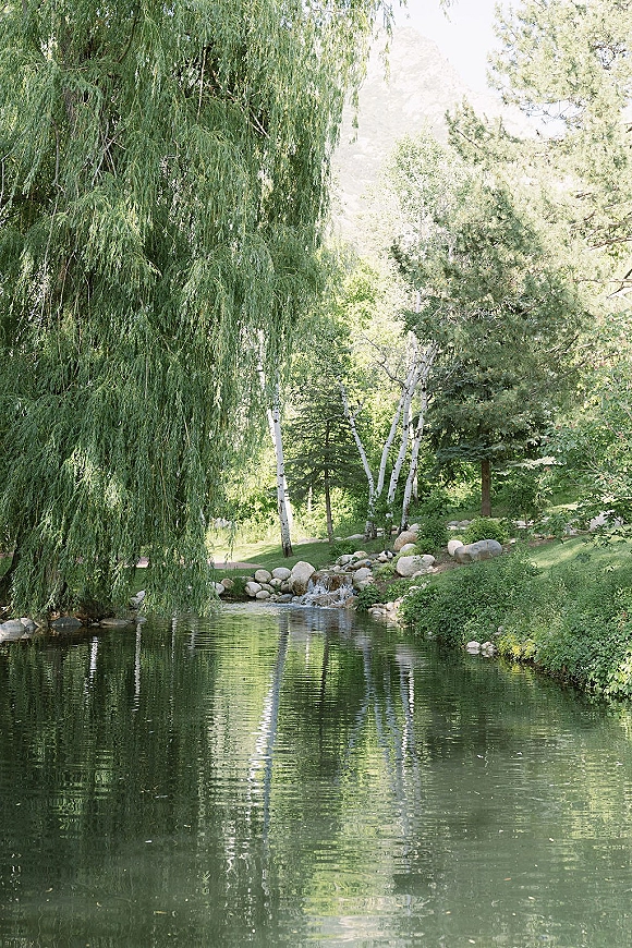 Outdoor wedding venue with a wedding venue pond, framed by a weeping willow, rocks, and mountain backdrop on a green lawn
