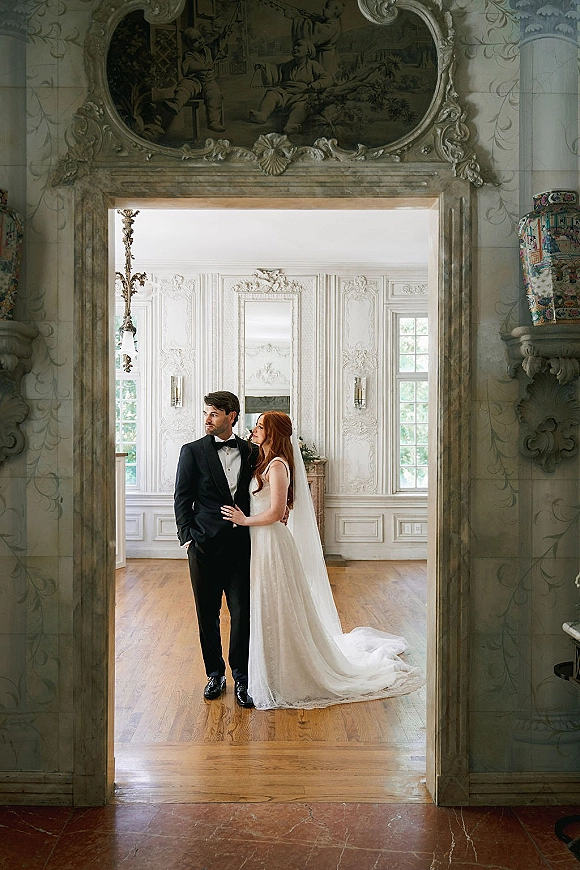 Couple portrait of bride in veil and wedding dress train beside groom in tuxedo, framed by an ornate doorway in a historic interior