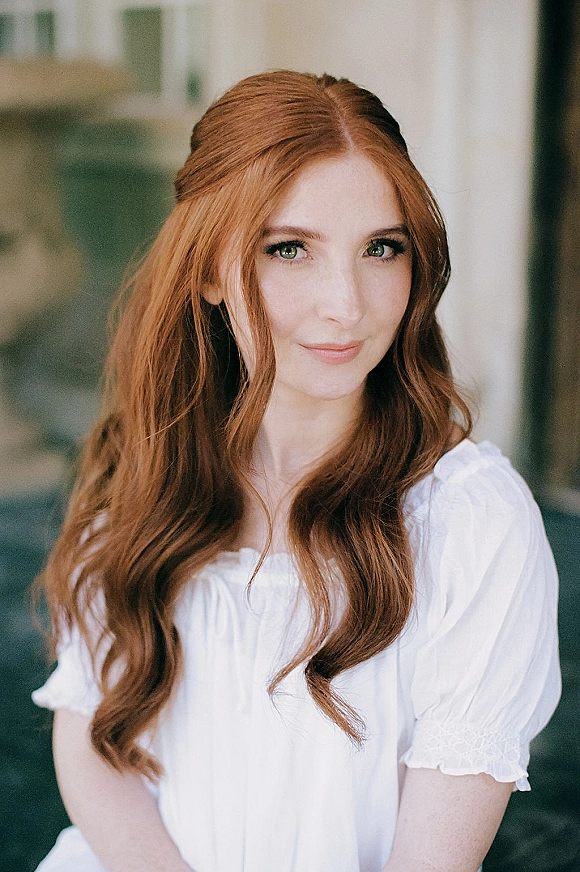 Bridal portrait of a redhead bride with long wavy half-up hair in a white puff-sleeve dress, softly lit by window light indoors