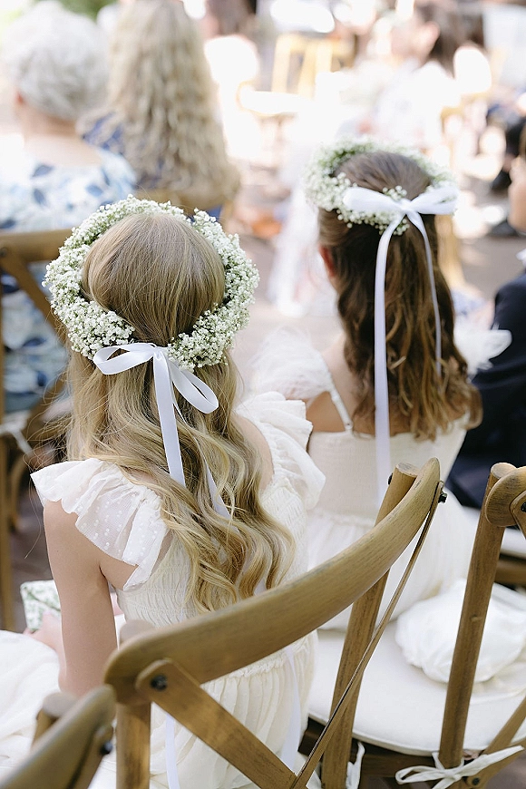Flower girl hairstyle with a baby's breath flower crown and white ribbon bow, back view in white dress near wooden ceremony chairs and guests