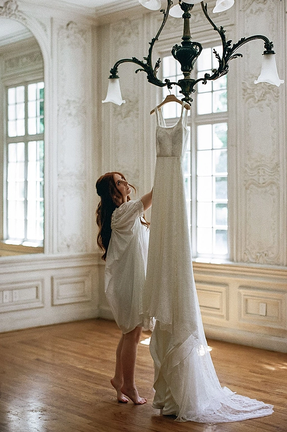 Bridal gown moment as a bride in a robe admires her wedding dress on a hanger beneath an ornate chandelier in natural light