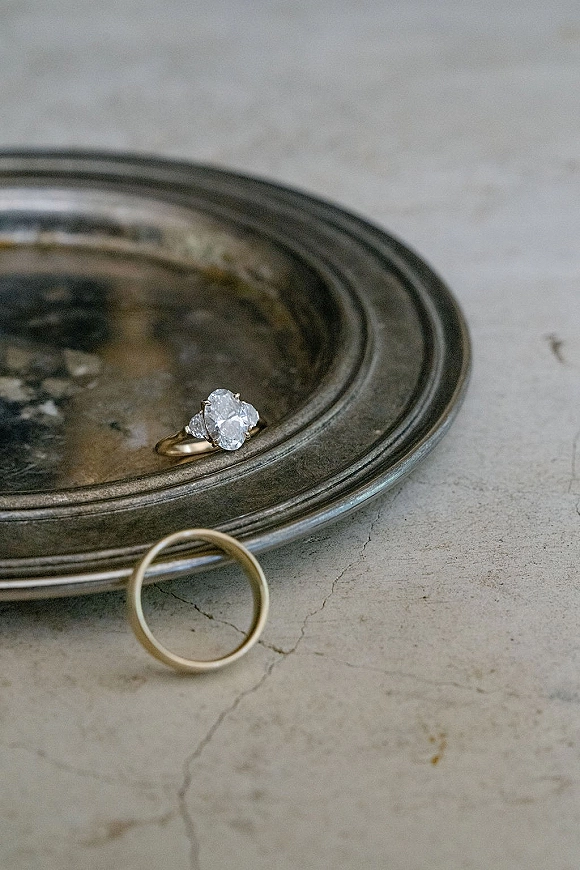 Wedding rings with an oval diamond engagement ring and thin gold band resting on a metal tray atop a neutral stone tabletop