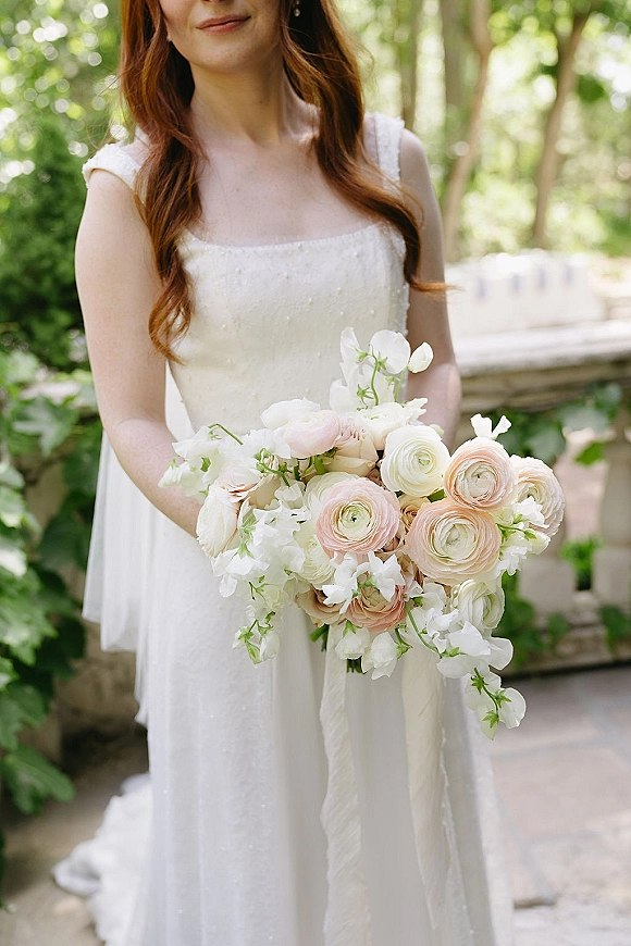 Bridal bouquet of blush and white ranunculus with sweet peas and a long silk ribbon, held by a bride in a lace dress in a garden