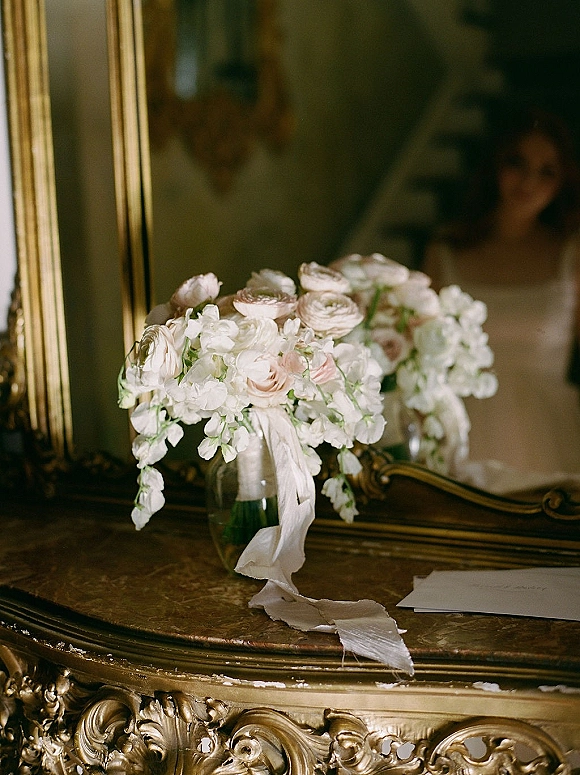 Bridal bouquet of white wedding bouquet blooms with blush accents and ribbon wrap in a glass vase on an ornate dresser by a gold-framed mirror