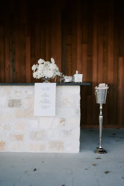 Wedding bar setup with a wedding bar menu sign, white rose centerpiece and silver tray on a stone bar front against a wood wall backdrop