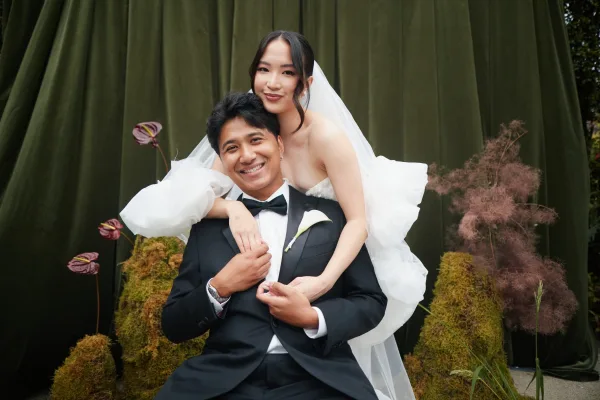 Couple portrait of bride hugging groom, highlighted by his calla lily boutonniere, against a green velvet drape and garden foliage.
