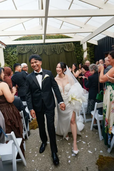 Wedding recessional with a just married exit as smiling newlyweds walk down the aisle, bride holding a white bouquet under a covered patio.