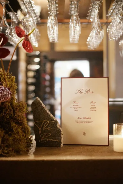 Wedding bar sign displaying a wedding drink menu, warmly accented by a single candle, set against a bar interior with a wooden ceiling.