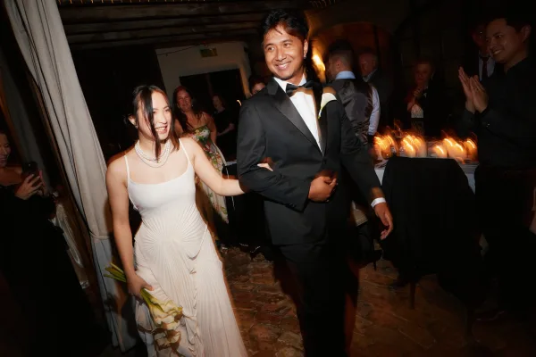 Reception entrance with newlyweds arm in arm, bride clutching a white calla lily bouquet beneath draped curtains by a candlelit table.