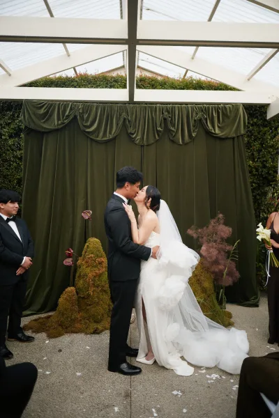 Wedding first kiss in a tender ceremony moment, the couple kissing under a pergola roof, framed by a rich velvet drape against a hedge wall.