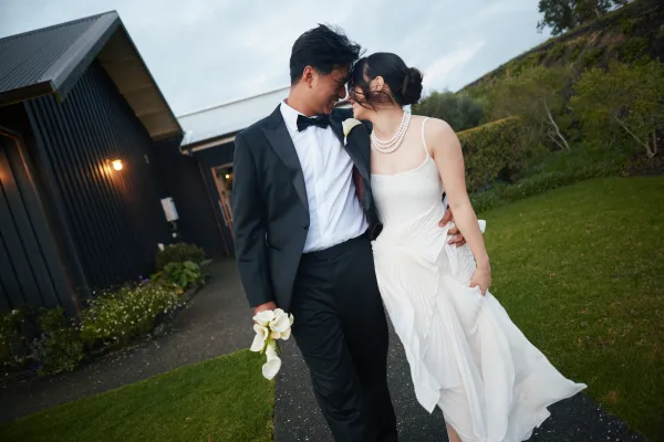 Couple portrait of the bride and groom walking along a garden walkway, her pearl necklace visible, with hedges and a building exterior behind them.