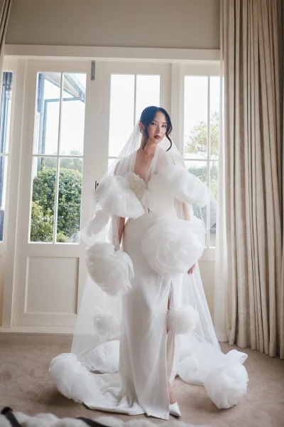 Bridal portrait of a bride in a 3D floral wedding dress, framed by French doors with garden trees, her long veil trailing over the carpet.