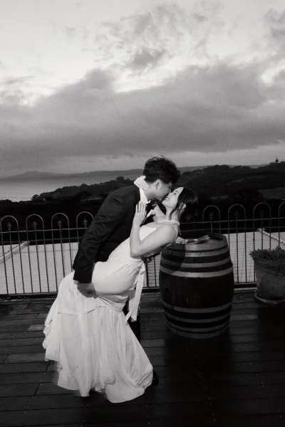 Wedding kiss portrait capturing a couple dip kiss, the groom dipping the bride on a terrace overlooking water and hills beside a wine barrel.