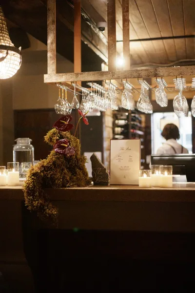 Wedding bar signage styled as a bar menu display beside a framed menu, set on a bar counter with wood beams and bar shelves behind.