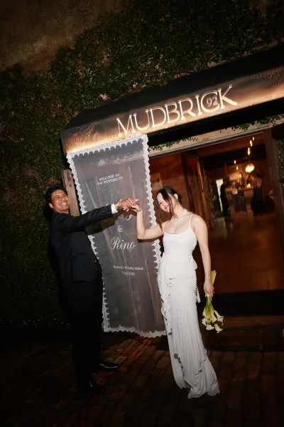 Couple portrait as the bride and groom twirl on a brick walkway by an ivy wall, her pearl necklace catching warm indoor lights.