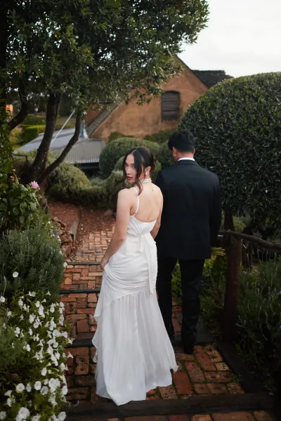 Couple portrait featuring an over-the-shoulder bridal portrait as they walk a brick garden path by a rustic building, her pearl necklace gleaming.