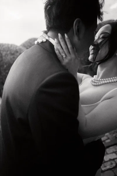 Wedding kiss portrait of a bride and groom close up, her engagement ring catching light as they embrace on a cobblestone path by garden hedges.