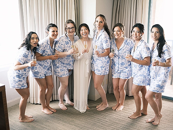 Bridesmaids getting ready in bridal party pajamas raise champagne flutes beside the bride in a white robe in a bright hotel room