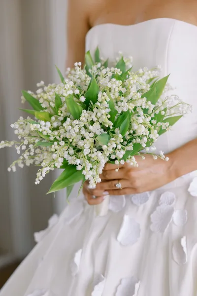 Bridal bouquet of lily of the valley held against a strapless gown with floral appliques, engagement ring visible by a neutral wall