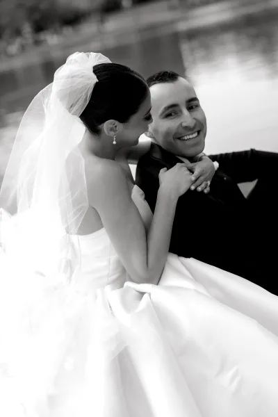 Couple portrait in a black and white wedding portrait style, bride in veil and strapless dress embracing smiling groom by the waterfront shoreline