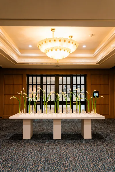 Reception escort table with wedding escort card display, calla lilies in glass cylinder vases and tea lights under a chandeliered ballroom ceiling