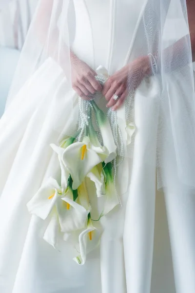Bridal bouquet of white calla lilies with pearl strands, held over a white dress and veil, with an emerald-cut ring in soft indoor light