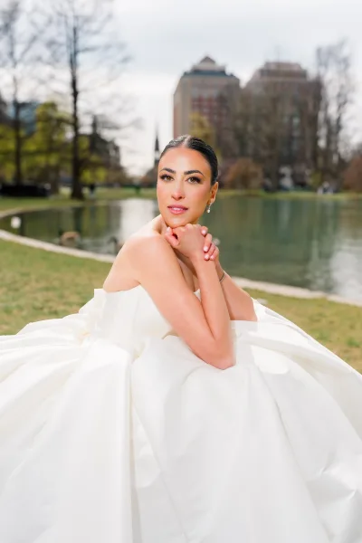 Bridal portrait of a bride in a strapless ball gown seated on grass, sleek low bun and drop earrings by a pond with city skyline