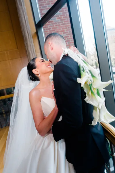 Wedding kiss portrait of bride and groom kissing by indoor windows, bride in strapless gown with veil holding calla lily bouquet against brick wall
