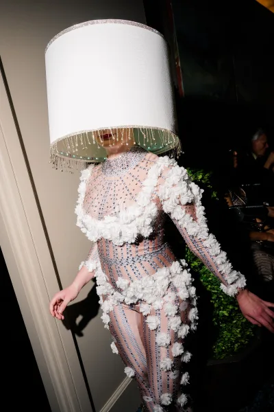 Bridal fashion look with a sheer beaded gown, 3D floral appliqués, and lampshade headpiece in a dim hallway with guests nearby