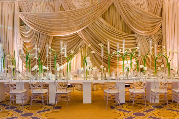 Reception tablescape with wedding head table decor, taper candles and white calla lilies in glass vases before a draped fabric backdrop in a ballroom with uplighting
