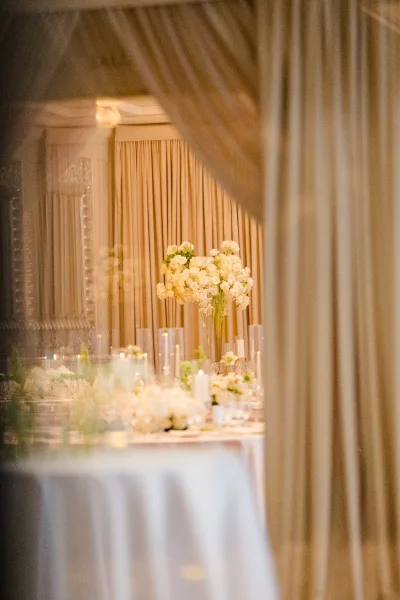 Reception tablescape with tall floral centerpiece in a clear glass vase, white blooms and candles glowing before draped curtains and a mirror reflection