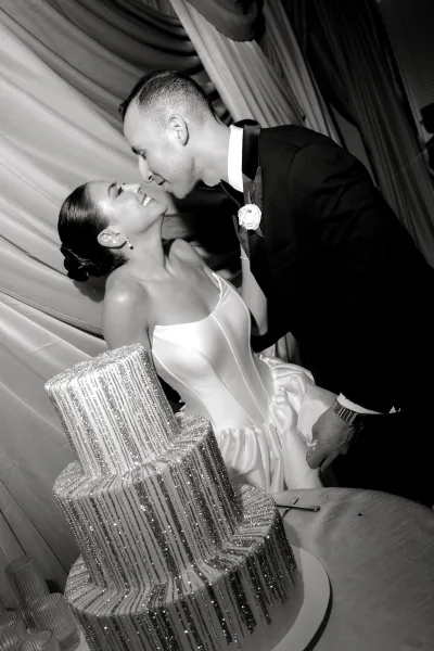 Wedding cake cutting as bride in strapless gown and groom in tuxedo hold the knife by a rhinestone cake against draped reception backdrop