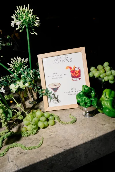 Signature drink sign for wedding signature cocktails in a frame on a marble tabletop with white flowers, greenery, grapes, and bell peppers against a dark backdrop