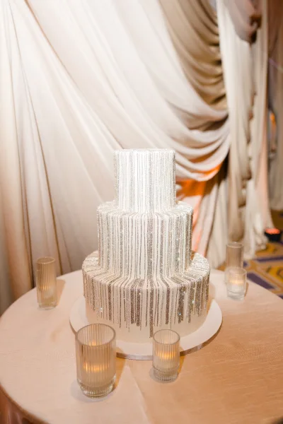 Wedding cake with silver beaded fringe on a round stand, surrounded by glass votive candles against a draped fabric backdrop indoors