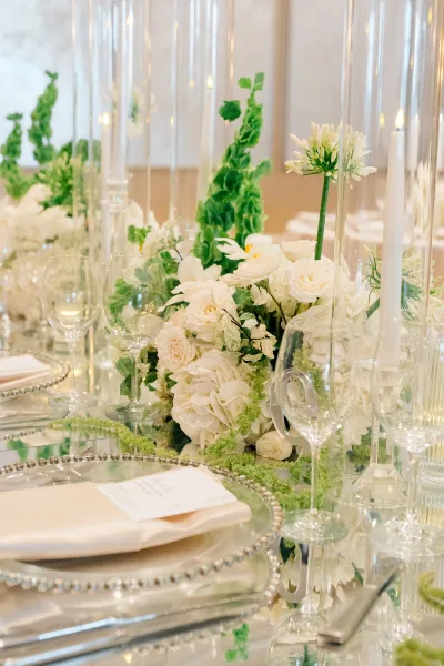 Reception tablescape with wedding table centerpiece of white flowers and greenery, hurricane vases and taper candles on a mirrored runner in a neutral room
