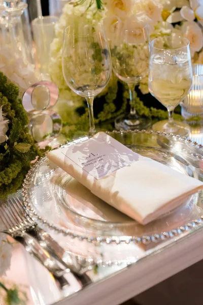 Reception tablescape with wedding place setting, clear glass charger, ivory napkin and menu card, rings by moss runner under soft lighting