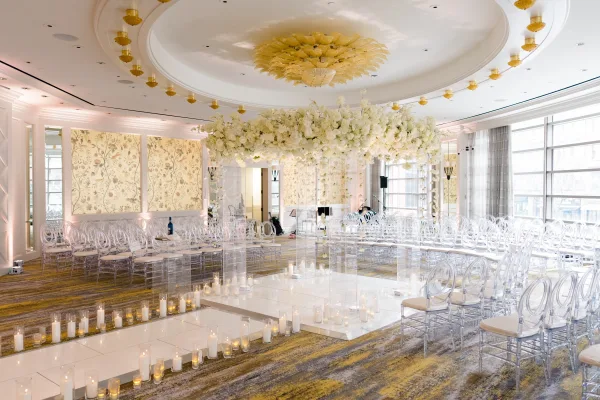 Indoor ceremony setup with wedding ceremony aisle candles, clear acrylic chairs, white runner, and hanging white florals in a windowed ballroom