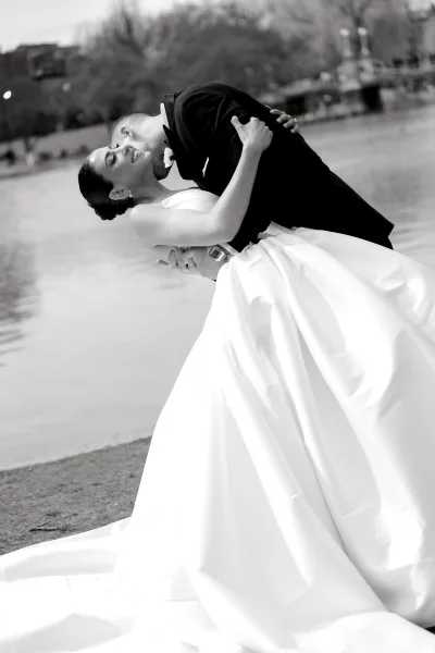 Wedding kiss portrait of groom dipping bride in a strapless dress with long train, beside lakeside water with trees and a bridge behind