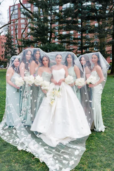 Bride with bridesmaids group photo showing a floral appliqué veil draped over them, holding white bouquets on a lawn with trees and apartments