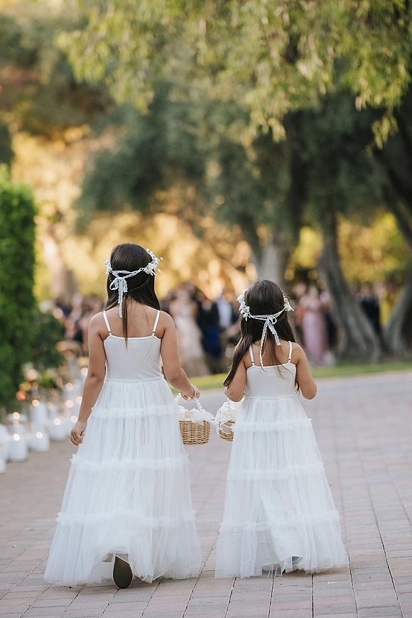 Flower girls in white tulle flower girl dresses with floral crowns and ribbon ties, holding baskets on an outdoor walkway among guests