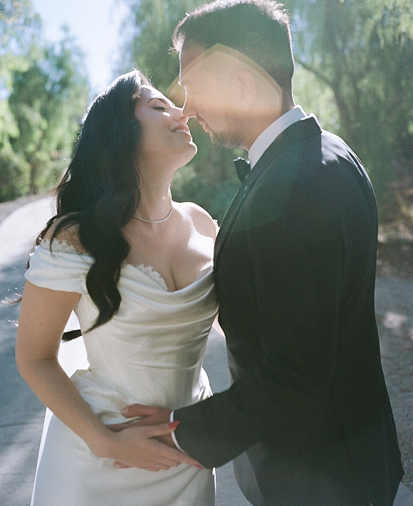 Wedding couple portrait of bride and groom in a wedding kiss moment, she in off-shoulder dress, backlit by sun flare on a tree-lined road