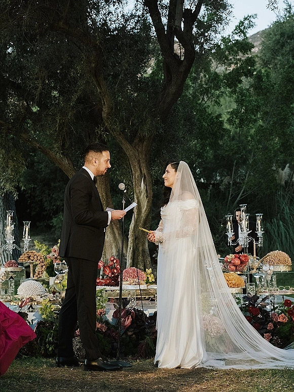 Wedding vows as groom reads vow cards beside bride in off the shoulder dress and long veil, at garden ceremony with candles and florals.