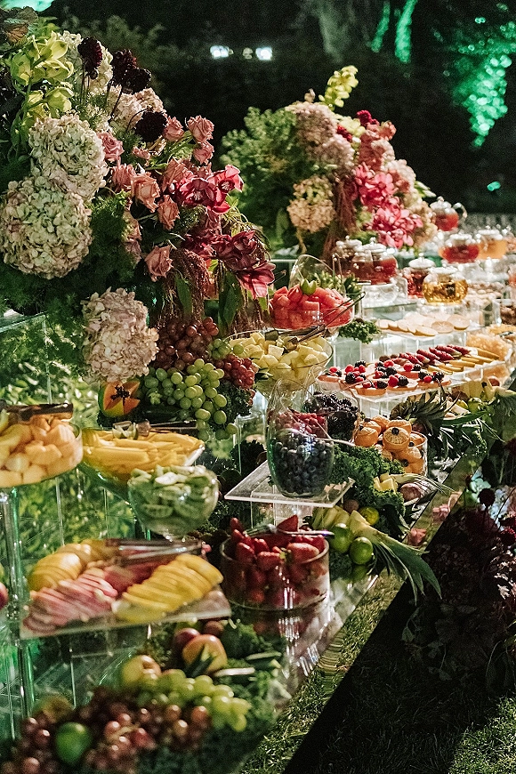 Wedding dessert table with wedding fruit display on glass risers, lush hydrangeas and roses, and sliced fruit and berry tarts outdoors at night