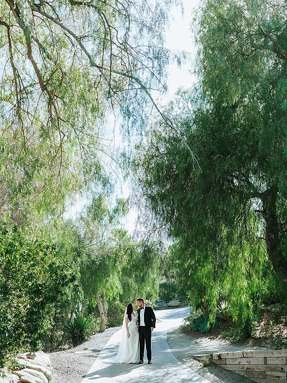 Couple portrait of bride in veil and groom in black tuxedo sharing a forehead kiss on a sunlit garden path beneath trees