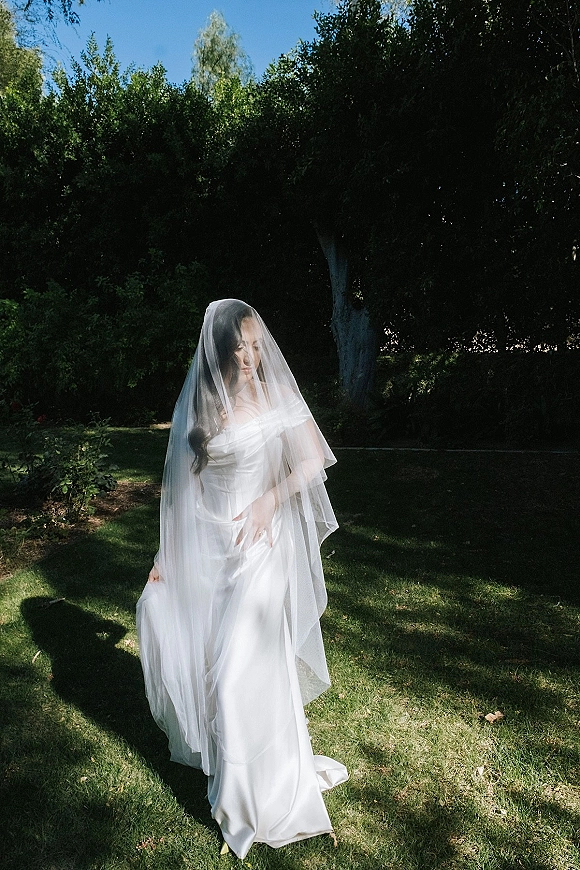 Bridal portrait of a bride in veil and off-the-shoulder wedding dress, standing on a sunlit garden lawn with trees and shadows